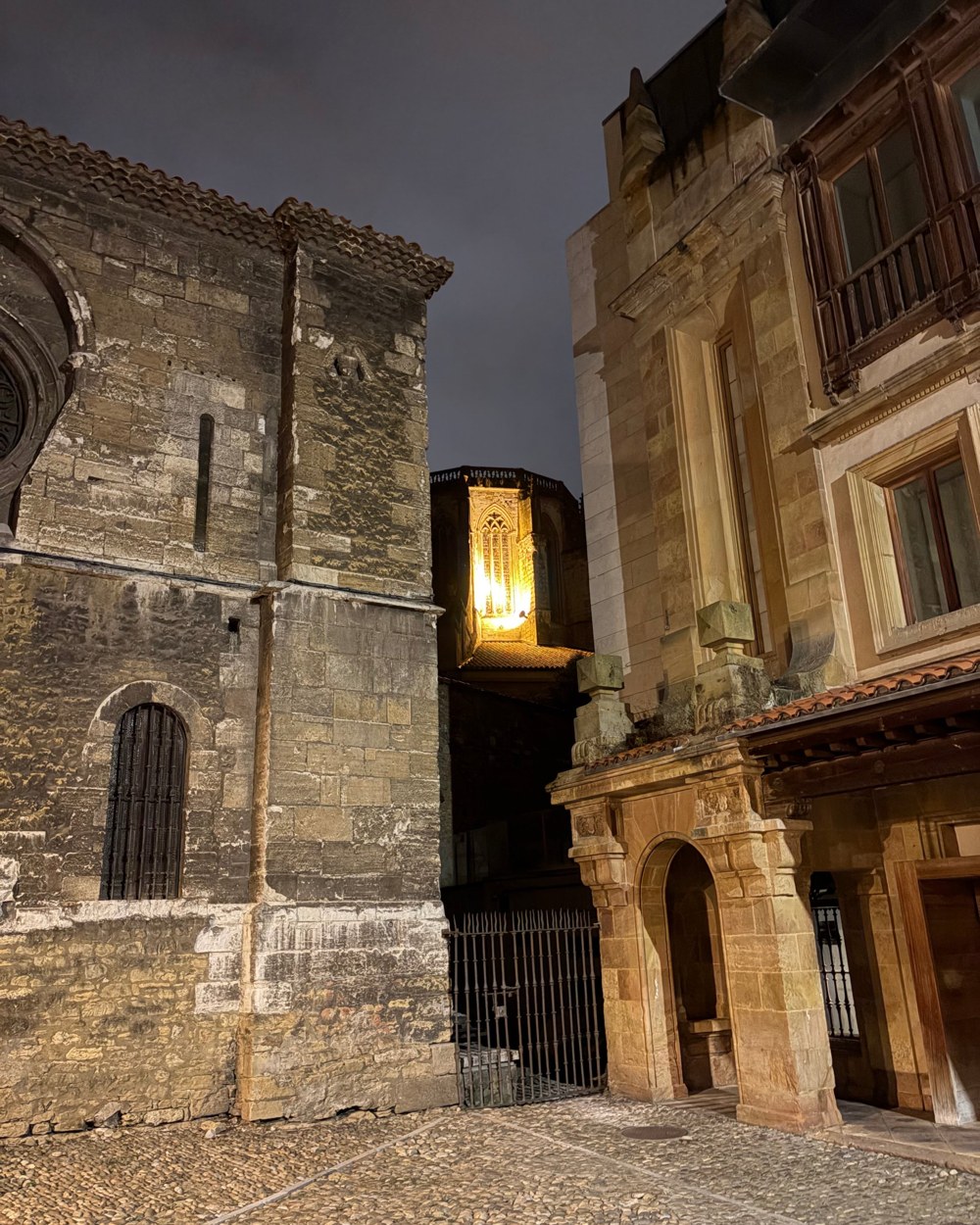 A narrow stone alley at night with an iron gate and a lit church tower behind